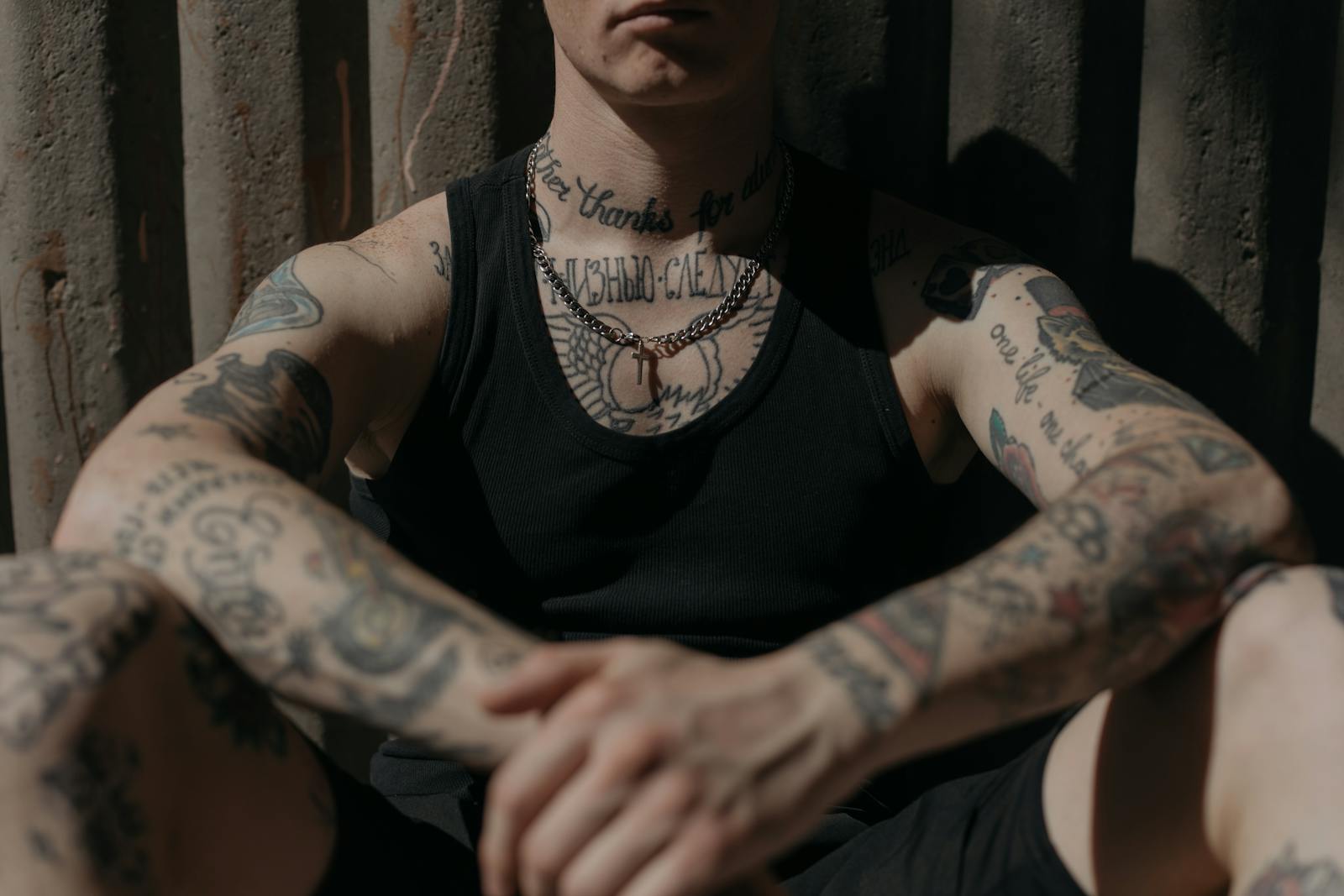 Close-up of a tattooed person in a black tank top sitting against a corrugated wall in moody lighting.